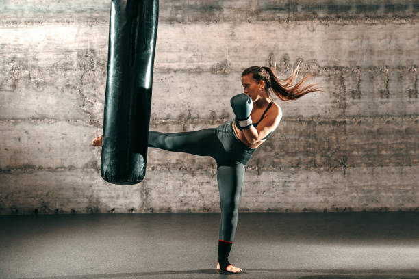 Dedicated strong brunette with ponytail, in sportswear, bare foot and with boxing gloves kicking sack in gym.